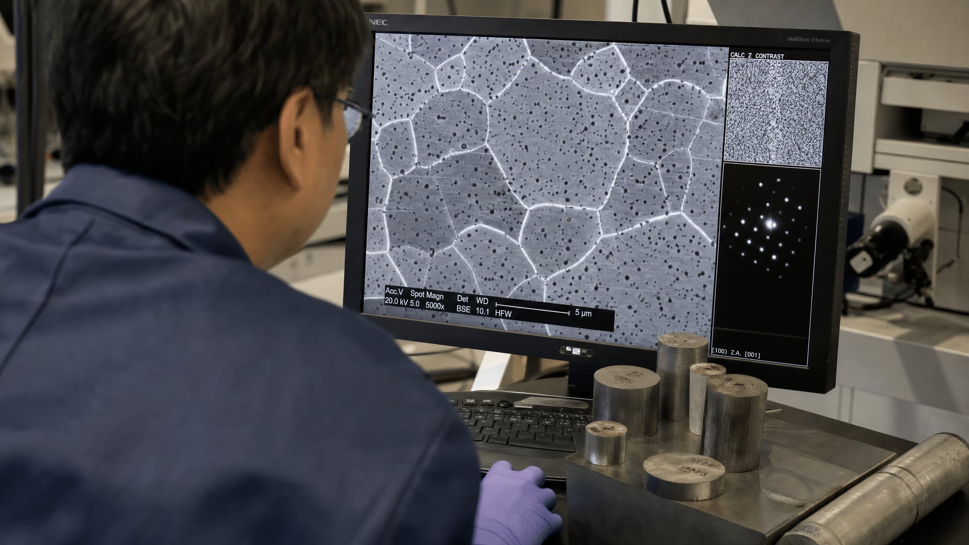 Metallurgist examining steel microstructure on an electron microscopy screen beside billet samples
